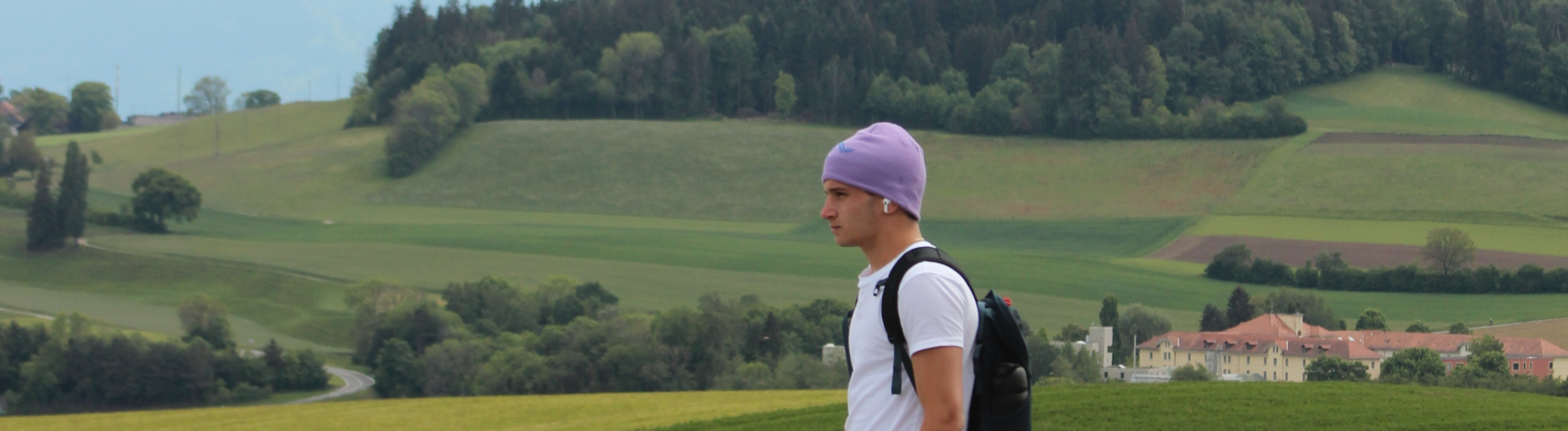 A man wearing a backpack and purple beanie walking through green Swiss countryside